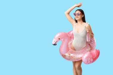 Mavi arka planda flamingo şeklinde şişme yüzük ve bir şişe kokteyl ile mayo giymiş güzel bir Asyalı kadın.