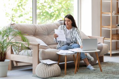 Genç ve güzel iş kadını elinde belgelerle kanepede oturuyor ve ofisteki bilgisayarını kullanıyor.