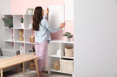 Oturma odasındaki pembe duvara boş tuval asan genç bir kadın.