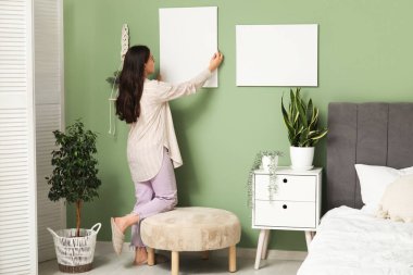 Yatak odasındaki yeşil duvara boş tuval asan genç bir kadın.