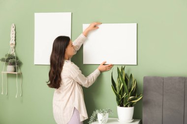 Yatak odasındaki yeşil duvara boş tuval asan genç bir kadın.