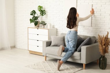 Oturma odasındaki beyaz tuğla duvara boş tuval asan genç bir kadın. Arka plan