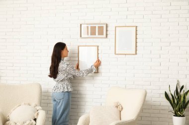 Evdeki beyaz tuğla duvara boş bir çerçeve asan genç kadın.
