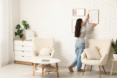 Evdeki beyaz tuğla duvara boş bir çerçeve asan genç bir kadın. Arka plan