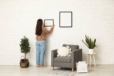 Evdeki beyaz tuğla duvara boş bir çerçeve asan genç bir kadın. Arka plan