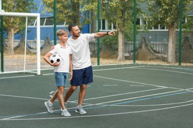 Babası ve oğlu futbol sahasında top oynuyorlar.
