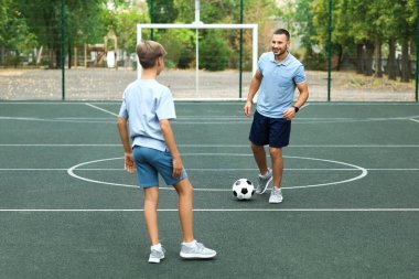 Çocuk babasıyla sahada futbol oynuyor.