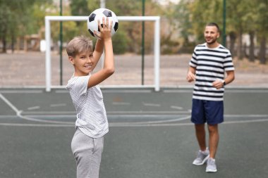 Çocuk babasıyla futbol sahasında futbol oynuyor.