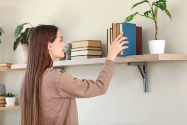 Oturma odasındaki raftan kitap alan genç bir kadın.