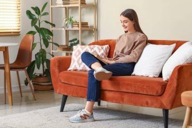 Oturma odasında kanepede oturmuş kitap okuyan genç bir kadın.