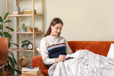 Oturma odasında kanepede oturmuş kitap okuyan genç bir kadın.