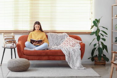 Genç bir kadın kanepede oturuyor ve oturma odasında kitap okuyor.