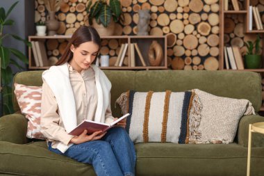 Oturma odasındaki kanepede oturmuş kitap okuyan güzel genç bir kadın.