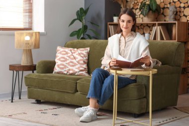 Oturma odasındaki kanepede otururken kahve masasında kitap okuyan güzel genç bir kadın.