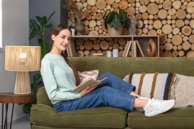 Oturma odasında kitapla kanepede oturan genç kadın.