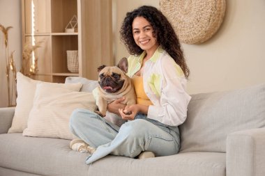Oturma odasındaki kanepede oturan sevimli Fransız bulldoguyla mutlu genç Afro-Amerikalı kadın.