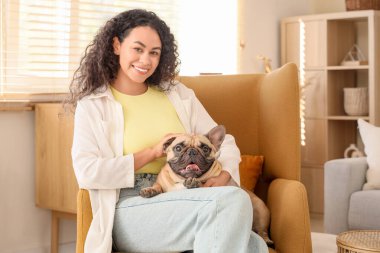 Evdeki koltukta oturan sevimli Fransız buldoguyla mutlu genç Afro-Amerikalı kadın.
