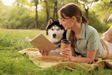 Bir fincan kahve ve parkta ekose üzerinde yatan sevimli Husky köpeğiyle kitap okuyan güzel genç bir kadın.