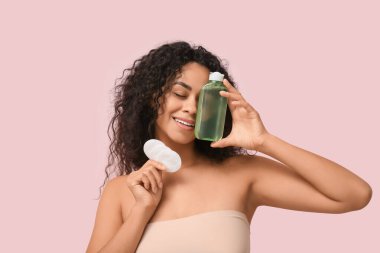 Beautiful young African-American woman with bottle of micellar water and cotton pads on pink background clipart