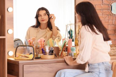 Güzel genç bir kadın evde aynanın yanında kaşlarını fırçalıyor.