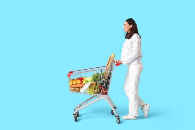 Mavi arka planda alışveriş arabası olan güzel, genç ve mutlu bir kadın.