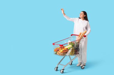 Mavi arka planda el sallayan alışveriş arabalı güzel, genç ve mutlu bir kadın.