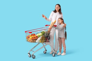 Güzel, genç, mutlu anne ve kızı ellerinde alışveriş arabalarıyla mavi arka plandaki bir şeyi işaret ediyorlar.