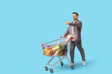 Alışveriş arabalı mutlu genç adam mavi arka planda bir şeyi işaret ediyor.