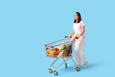 Mavi arka planda alışveriş arabası olan güzel, genç ve mutlu bir kadın.