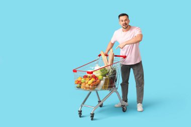 Şok olmuş genç adam mavi arka plandaki alışveriş arabasını işaret ediyor.