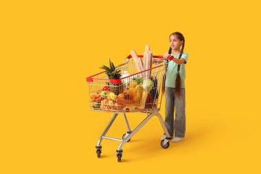 Sarı arka planda alışveriş arabası olan şirin, mutlu küçük kız.