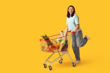 Sarı arka planda alışveriş arabası olan güzel, genç ve mutlu bir kadın.