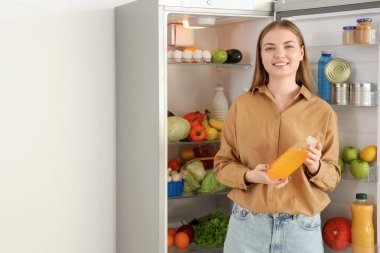 Mutfaktaki açık buzdolabının yanında meyve suyu şişesi olan genç bir kadın.