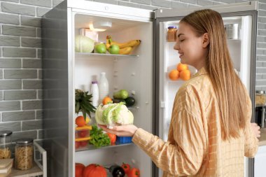 Mutfaktaki açık buzdolabının yanında lahana taşıyan genç bir kadın.