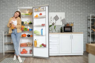 Mutfaktaki açık buzdolabının yanında meyve suyu içen genç bir kadın cep telefonuyla konuşuyor.