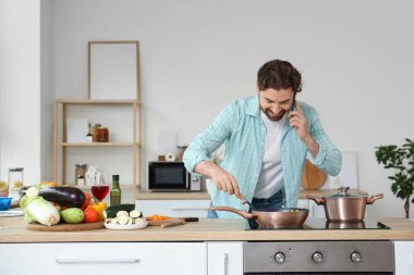 Yakışıklı sakallı adam mutfakta fırında sebzeyle cep telefonuyla konuşuyor.