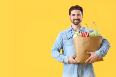 Sarı arka planda yiyecek dolu çantası olan yakışıklı sakallı adam.