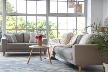 Oturma odasının içinde gri koltuklar ve vazoda şakayık çiçekleri var.