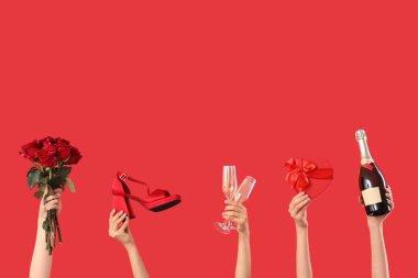 Kalp şeklinde hediye kutusu, güller, topuklu ayakkabılar, kırmızı arka planda şampanya şişesi olan kadın elleri. Sevgililer Günü kutlaması