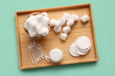 Wooden tray with cotton balls, pads, swabs and jar of cosmetic cream on turquoise background clipart