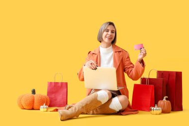 Sonbahar elbiseli genç bir kadın dizüstü bilgisayarı ve kredi kartı ile sarı arka planda online alışveriş yapıyor.