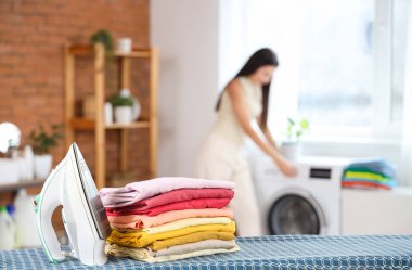 Demiri ve giysi yığınıyla güzel genç bir kadın banyoda çamaşır makinesini açıyor.