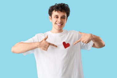 Yakışıklı genç adam mavi arka planda kağıt kalbe işaret ediyor.