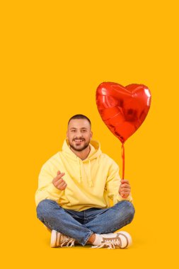 Kalp şeklinde balonu olan yakışıklı genç adam kalp hareketi gösteriyor ve sarı arka planda oturuyor.