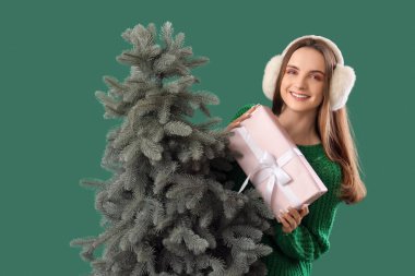 Yeşil arka planda, Noel ağacının yanında hediye kutusuyla şık, sıcak giysiler içinde güzel bir genç kadın.