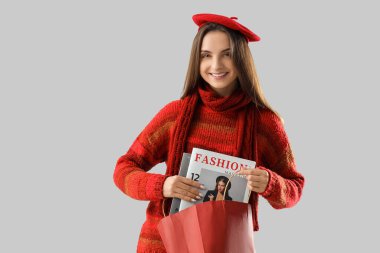 Gri arka planda, alışveriş çantası ve dergilerle şık ve sıcak kıyafetler içinde güzel bir genç kadın.