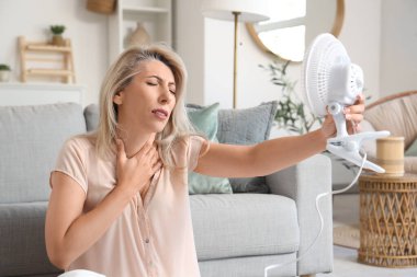 Mature woman with electric fan suffering from hot flashes at home. Menopause concept clipart