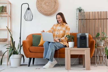 Evinde taşınabilir elektrik santrali olan koltukta dizüstü bilgisayar kullanan genç bir kadın.