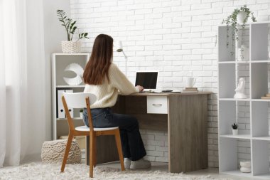 Modern dizüstü bilgisayarla çalışan genç bir kadın odasındaki beyaz tuğla duvarın yanındaki masada.