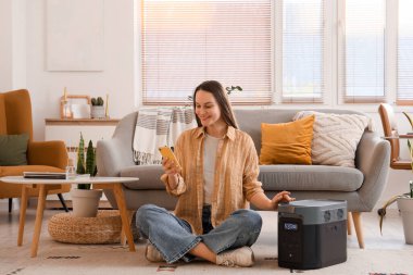 Evdeki halı üzerinde taşınabilir elektrik santrali olan cep telefonu kullanan genç bir kadın.